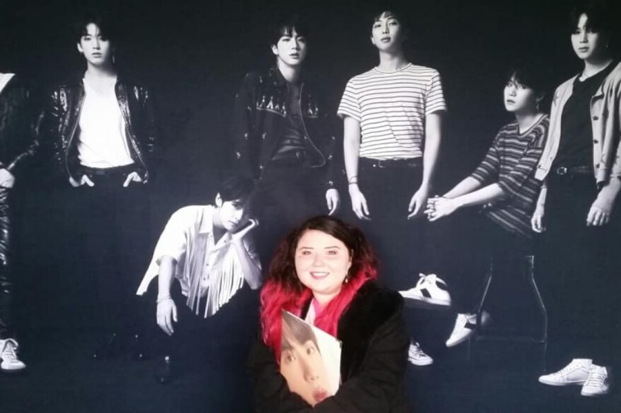 Cartoonist Emily Hacker poses with a backdrop featuring KPOP superstars BTS.